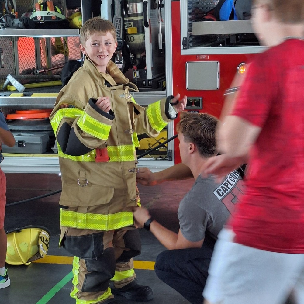 Child wearing an oversized firefighter suit gets help fastening the coat in front of a fire engine